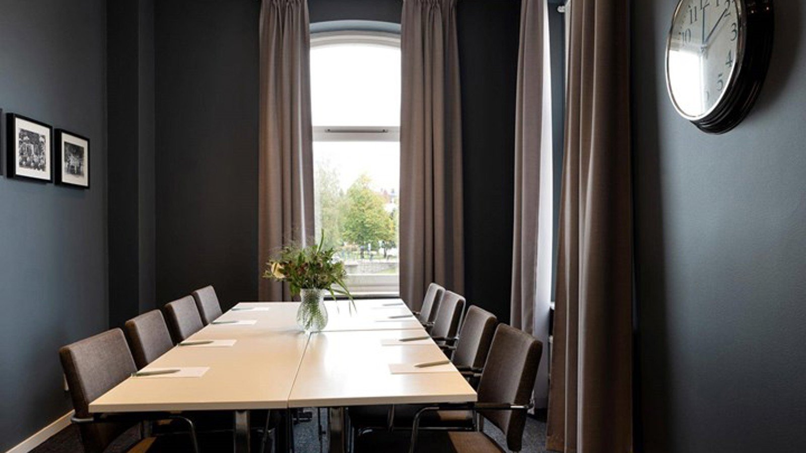 Konferensrum med styrelsesittning, stora fönster, mörka väggar, vitt bord med blomvas