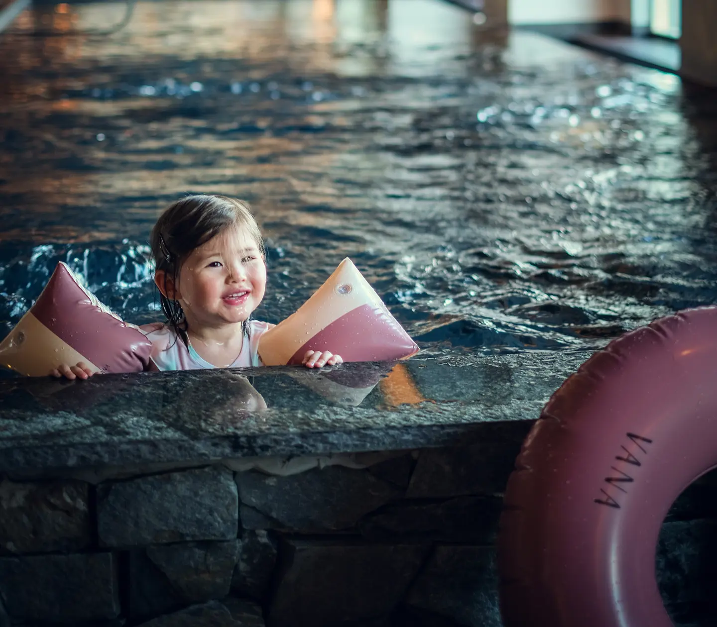 Litet barn med flytkuddar badar i en inomhuspool och ser glatt ut