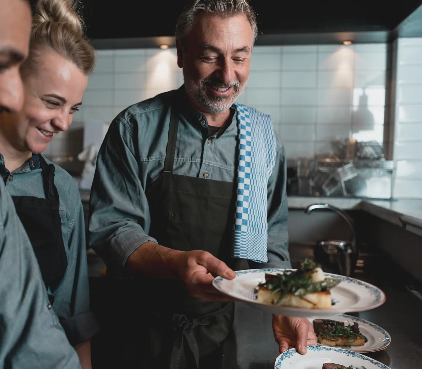 Chef presenting plated dishes to kitchen staff in a modern restaurant kitchen, showcasing teamwork and culinary presentation skills.