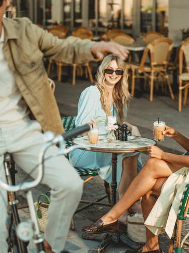 Unga vänner som njuter av fika utomhus på ett kafé i staden, man på cykel hälsar på sittande kvinnliga vänner vid bordet.