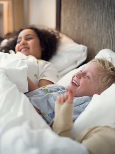 Tre barn i pyjamas som ligger i hotellsäng och skrattar