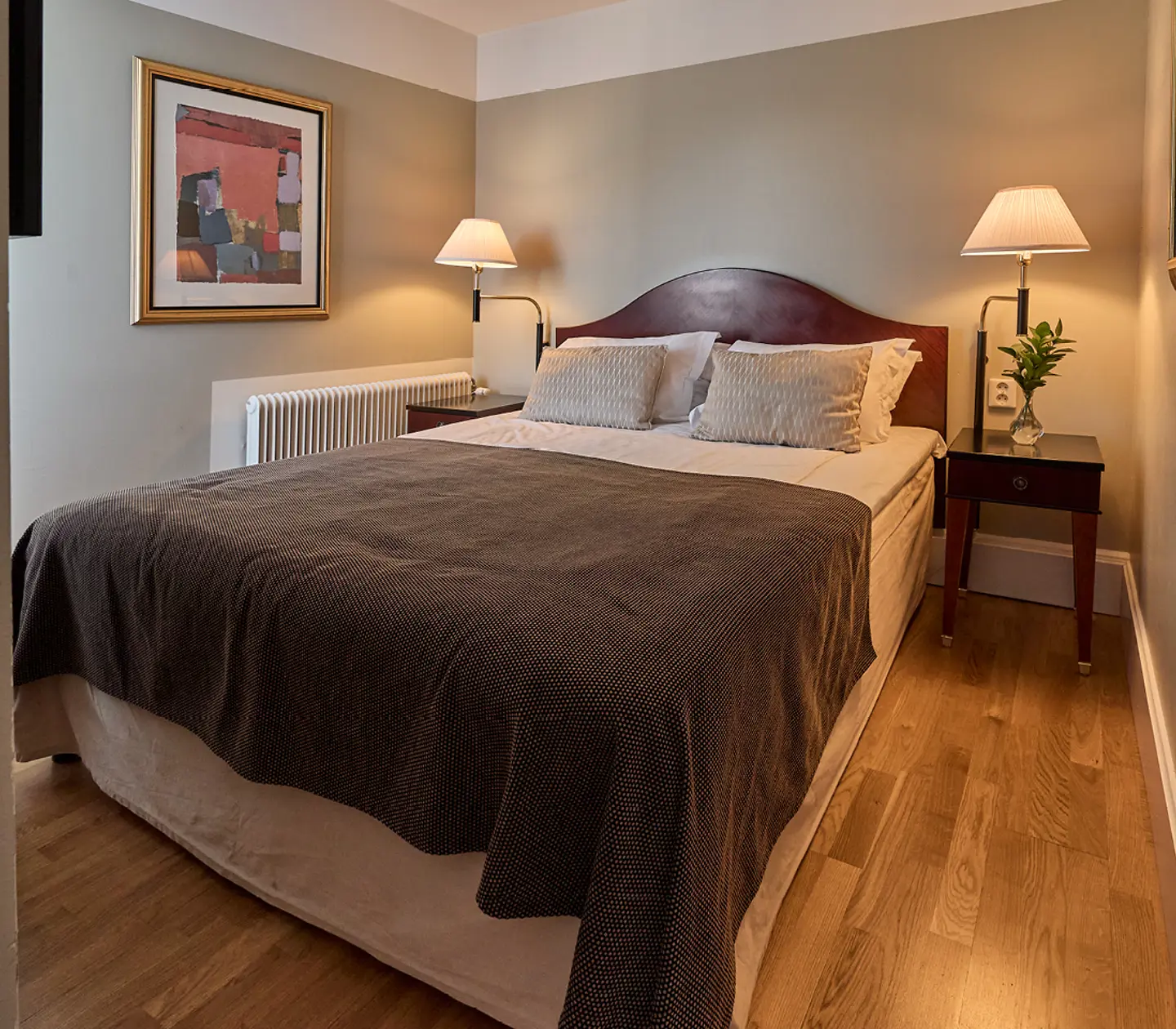 Cozy bedroom with a neatly made bed, wooden nightstands, and warm lighting, featuring modern artwork and a hardwood floor.