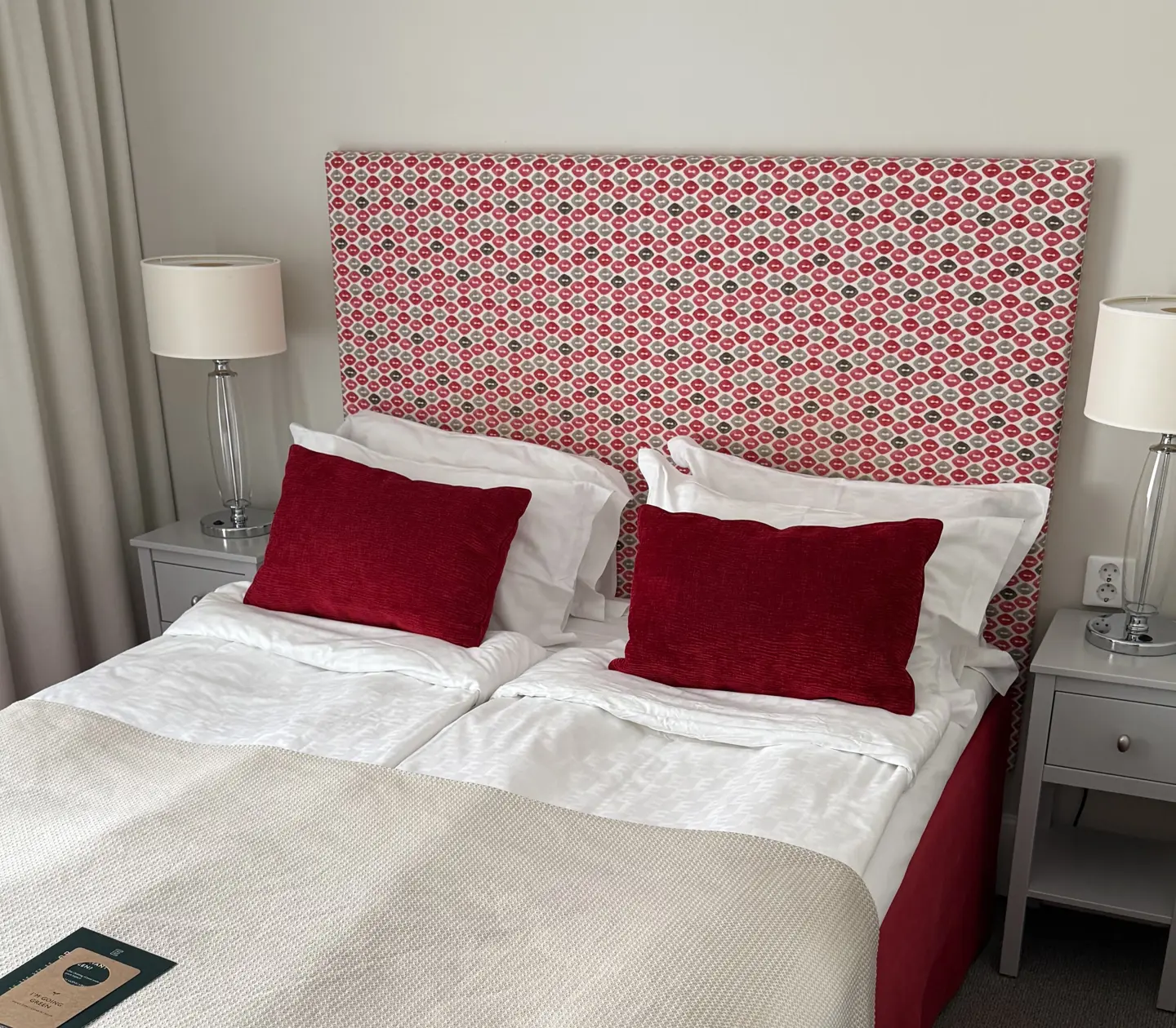 Cozy hotel room with twin beds featuring red patterned headboards, white bedding, and red accent pillows. Modern bedside tables with glass lamps add elegance.
