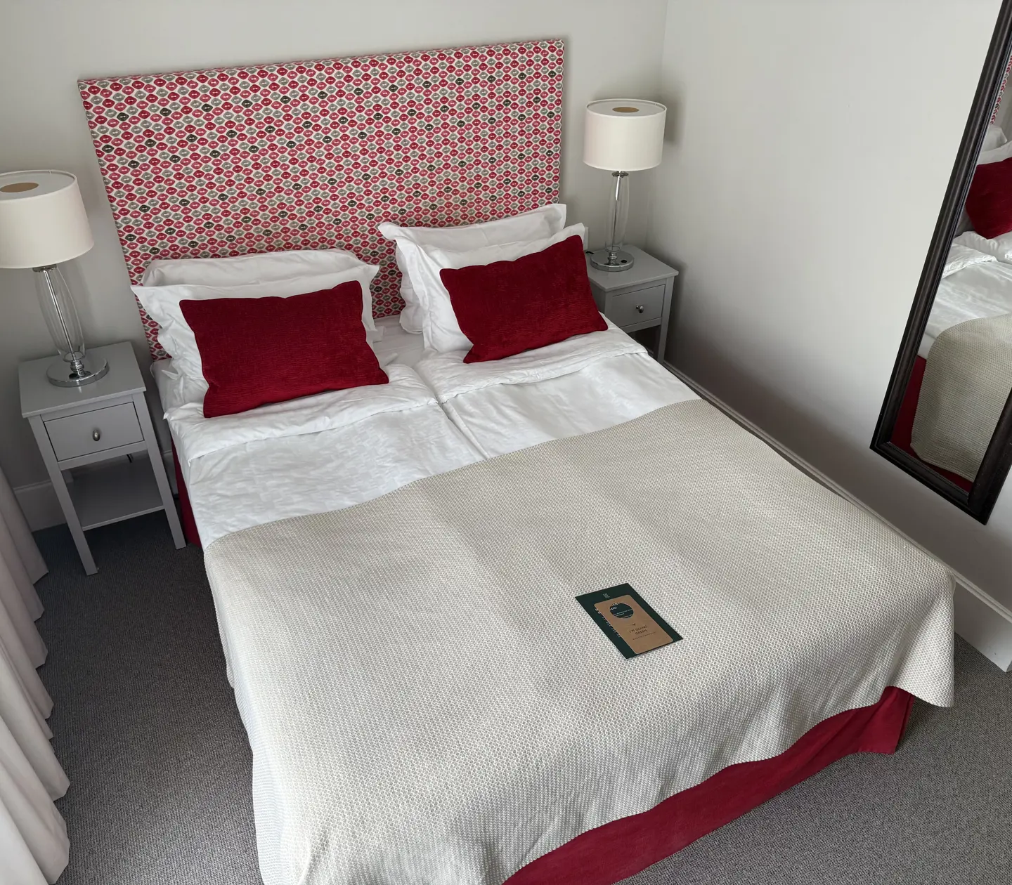 Cozy hotel room featuring a double bed with a decorative floral headboard, red accent pillows, and a beige blanket, accompanied by bedside tables with modern lamps.