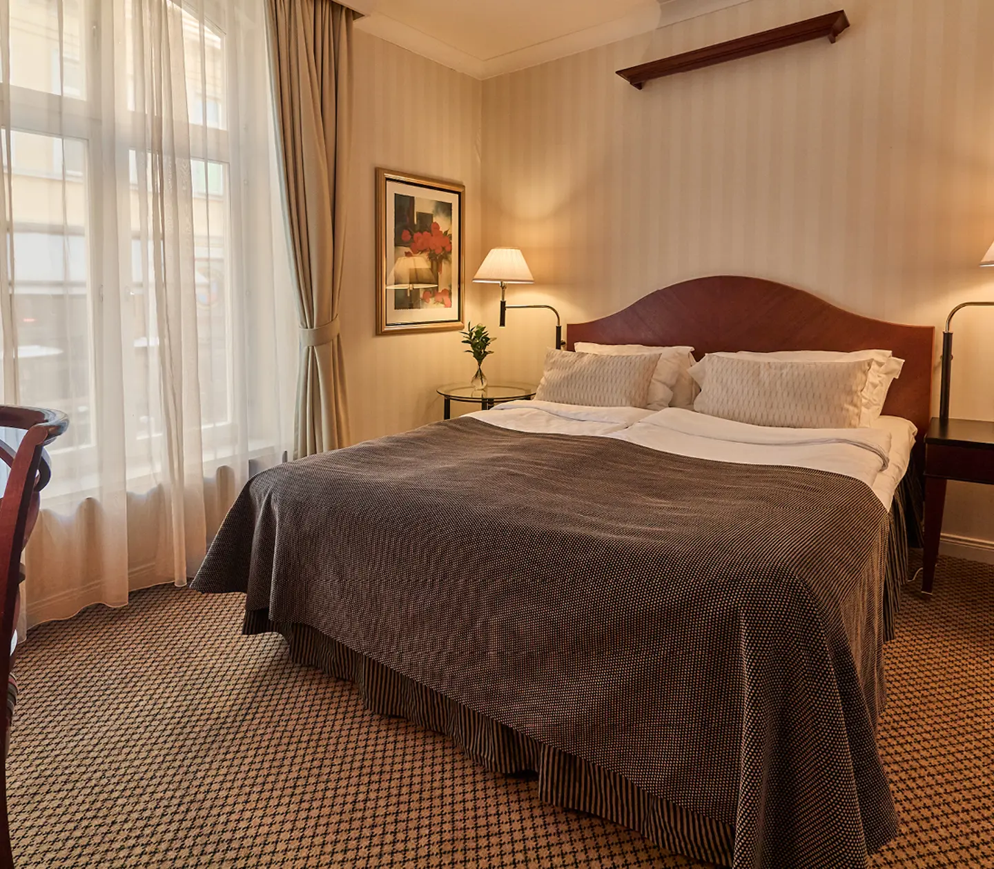 Elegant hotel room with a cozy double bed, two armchairs, bedside tables, and large curtained windows, providing a comfortable and stylish ambiance for guests.