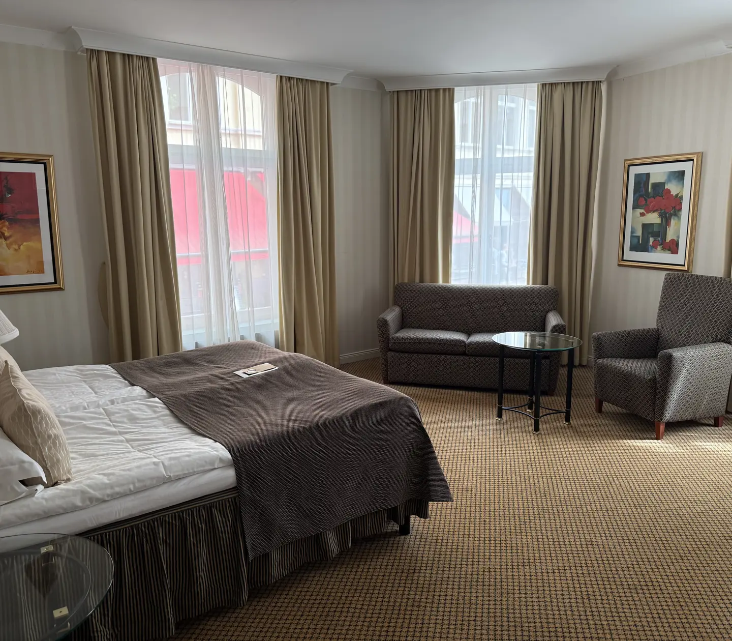 Modern hotel room with a cozy bed, stylish gray armchair, comfortable sofa, and round glass coffee table, accented by framed art and large windows with beige curtains.