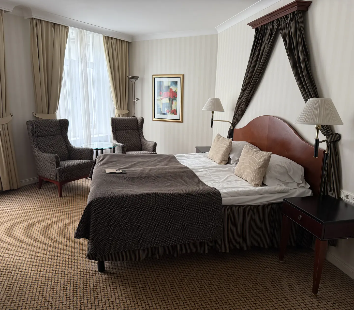 Elegant hotel room with a cozy double bed, two armchairs, bedside tables, and large curtained windows, providing a comfortable and stylish ambiance for guests.