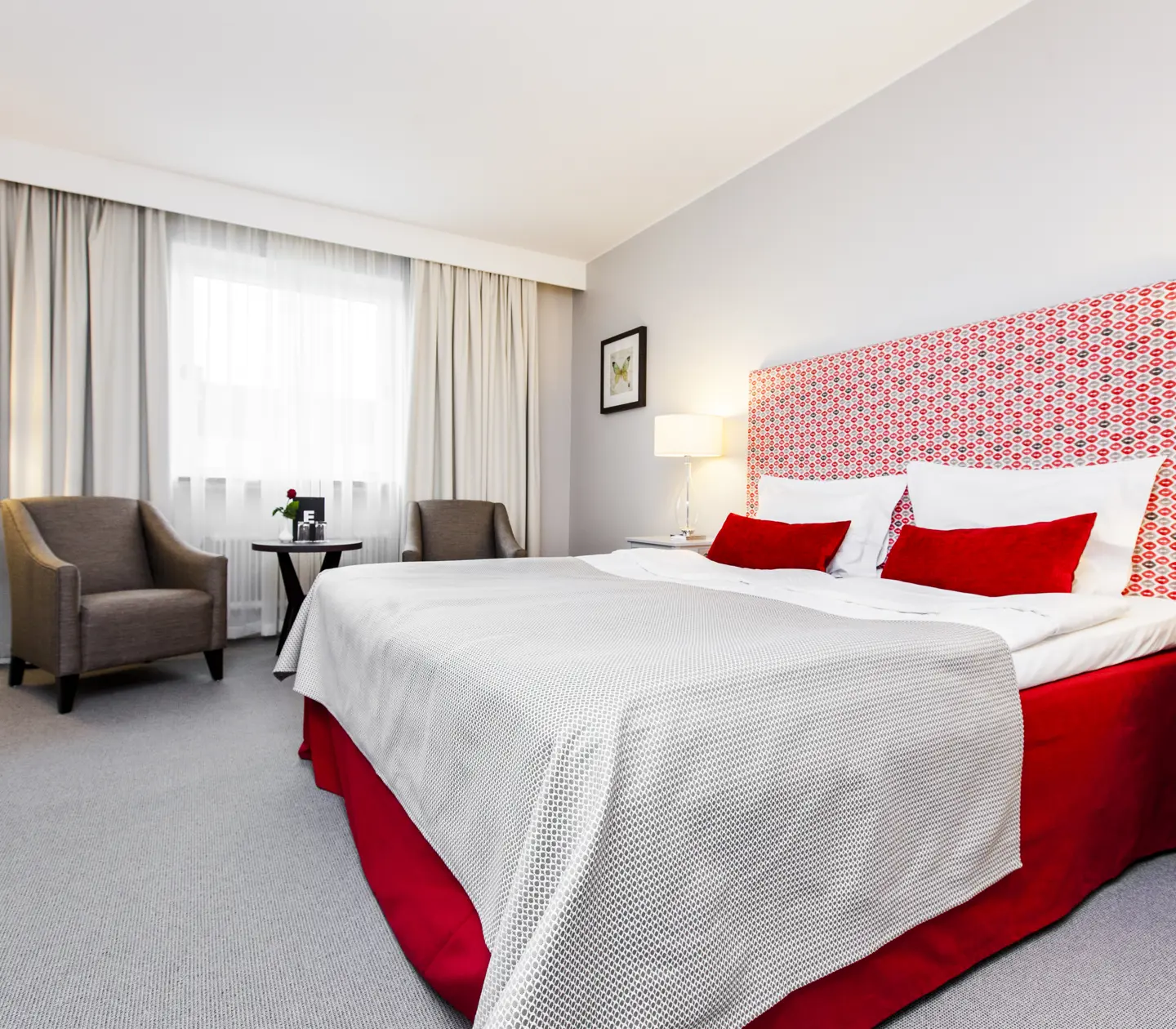 Bright hotel room with bed, red headboard and armchairs