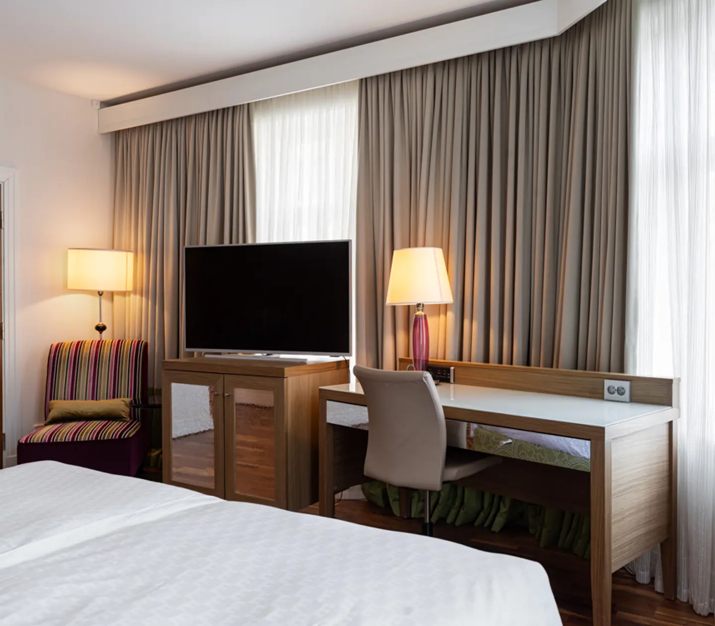 Modernt hotellrum med en stor säng, skrivbord med stol och platt-TV. Fönster med eleganta gardiner släpper in naturligt ljus.