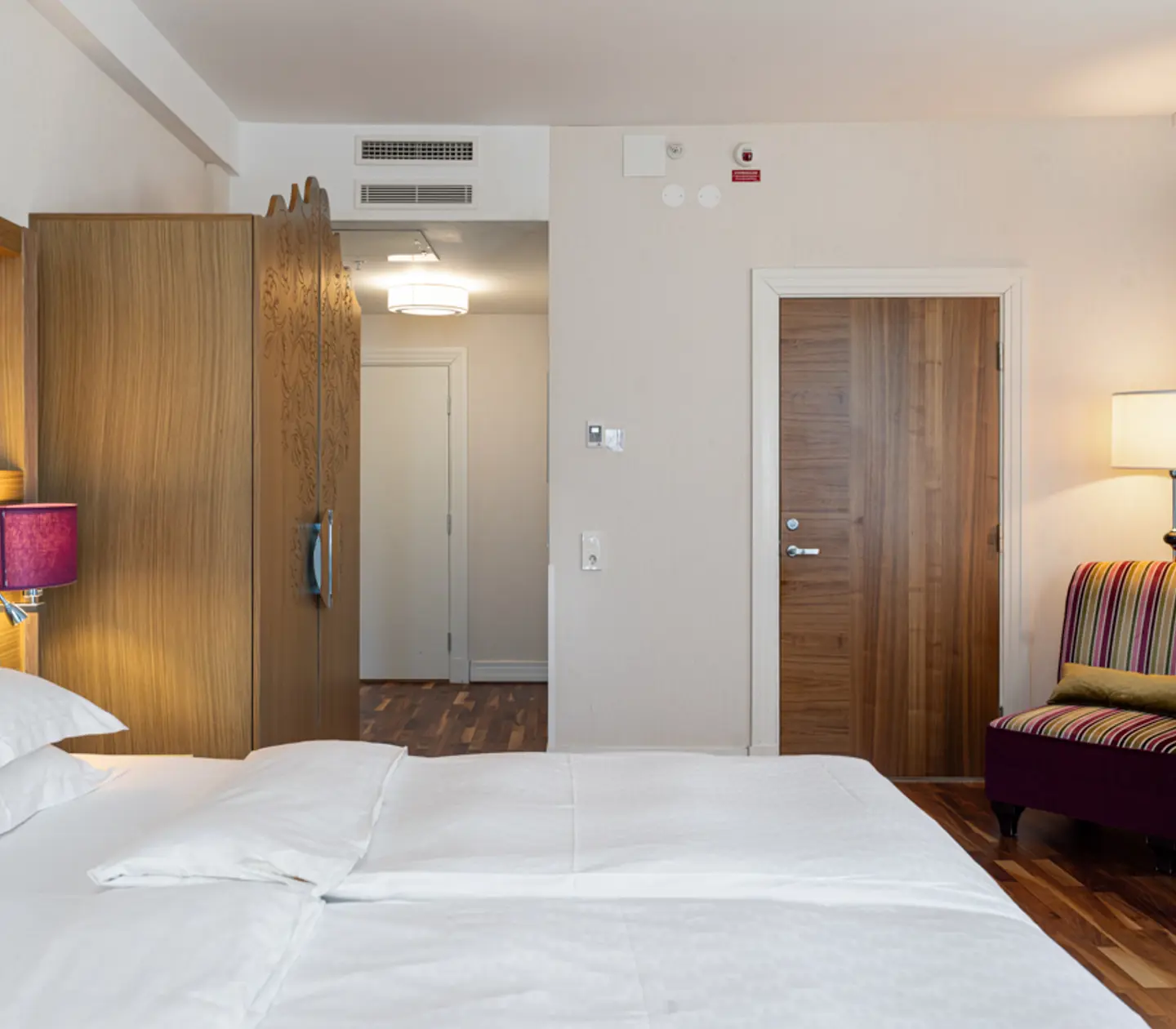 Modernt hotellrum med dubbelsäng, trädetaljer och en randig fåtölj bredvid en golvlampa.