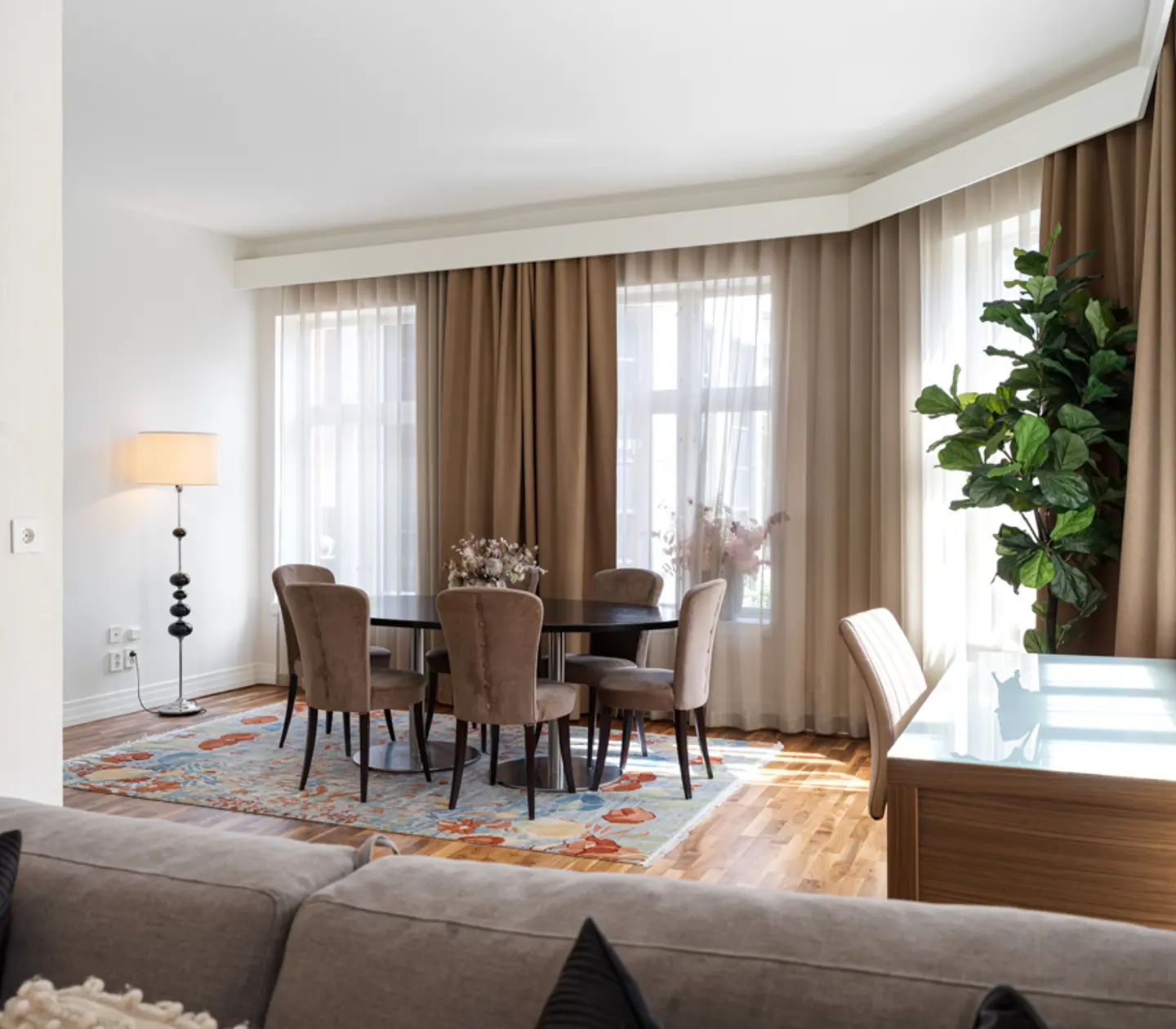 Ljust och luftigt vardagsrum med matsalsbord och stolar, bredvid stora fönster med beige gardiner. Gröna växter och en golvlampa kompletterar den eleganta inredningen.