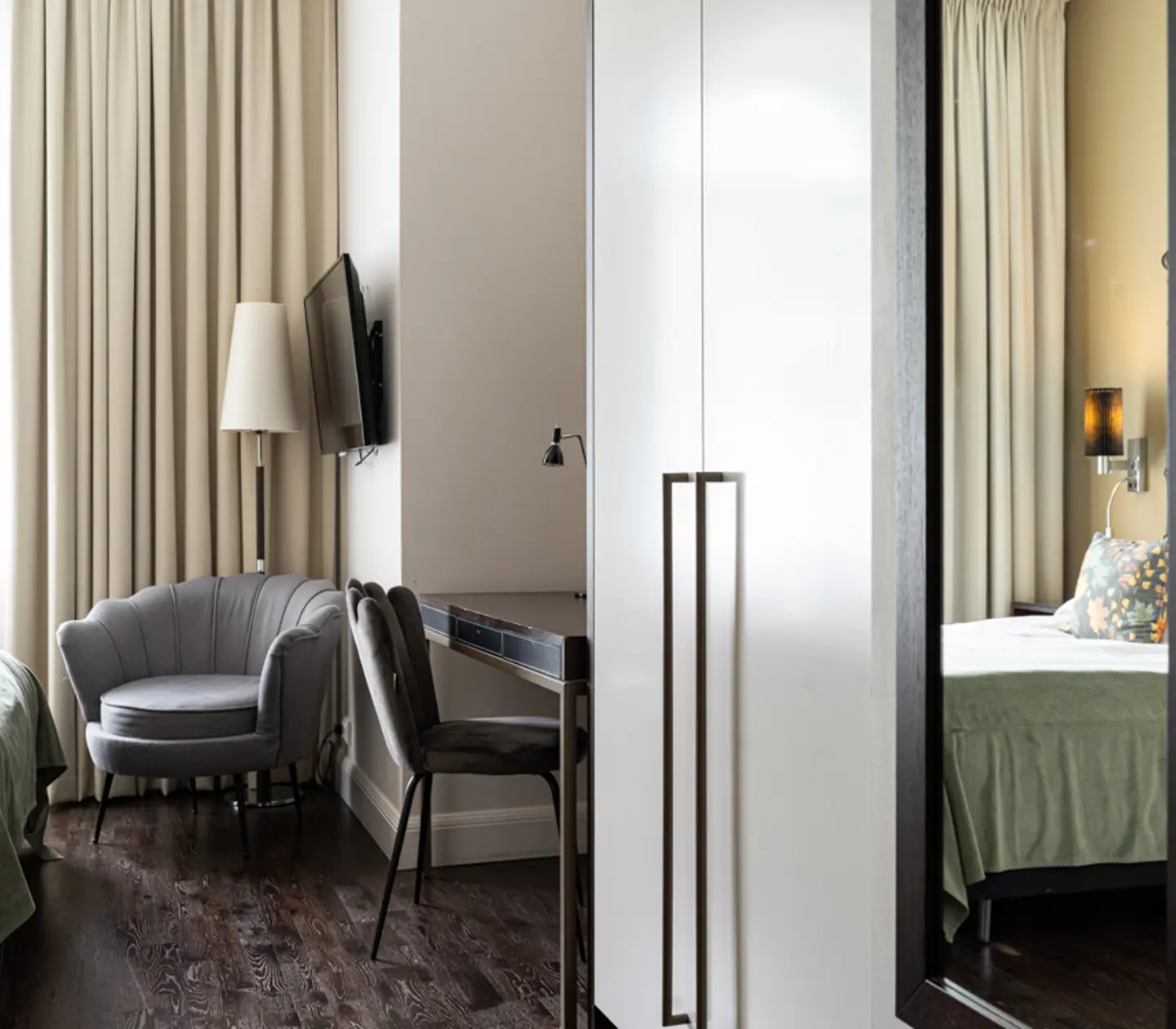 Modern hotel room interior with sleek design features a cozy gray armchair, a mounted flat-screen TV, a work desk with chairs, and a comfortable bed. Elegant beige curtains and dark wood flooring enhance the room's sophisticated aesthetic.