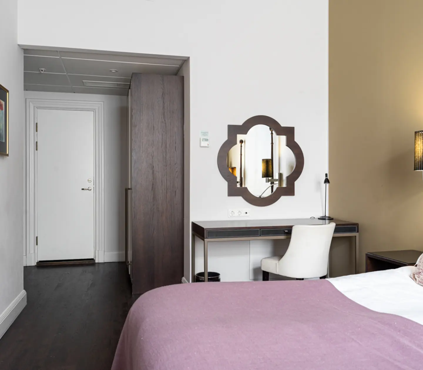 Hotel room interior featuring a bed with a purple cover, a stylish wall mirror above a workstation, a modern chair, decorative wall lamps, and a muted color palette for a cozy atmosphere.