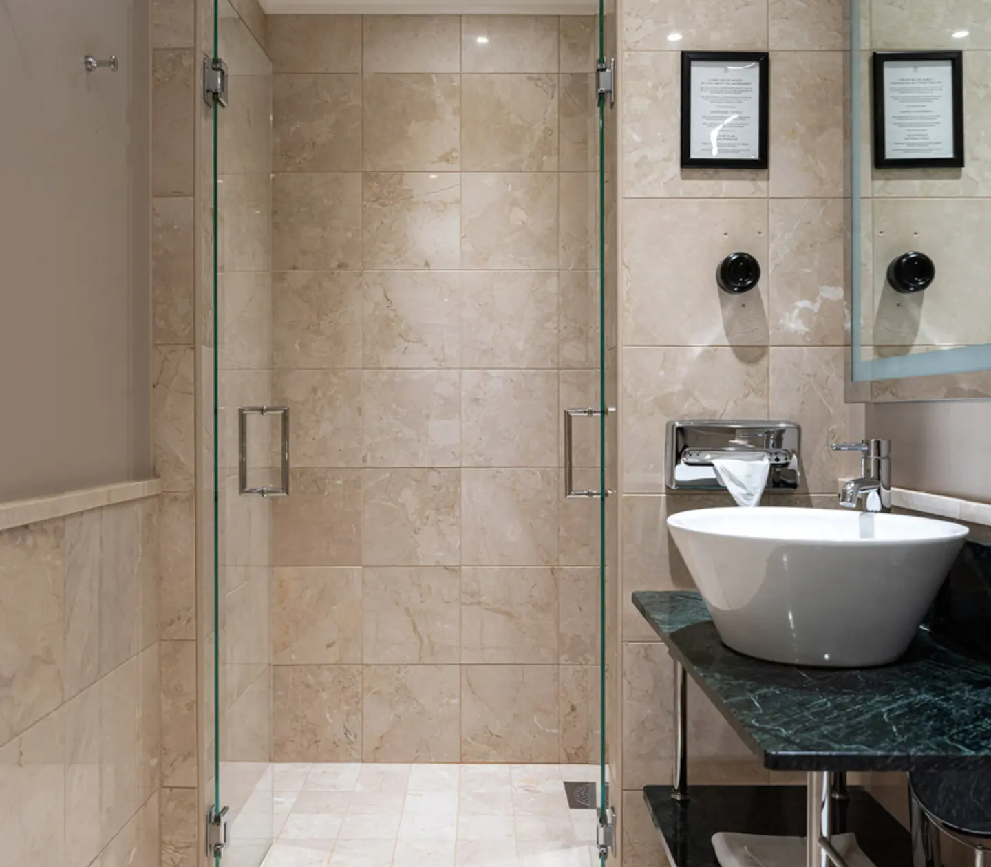 Modern bathroom with glass-enclosed marble-tiled shower, white sink on dark countertop with toiletries, and framed wall decor.