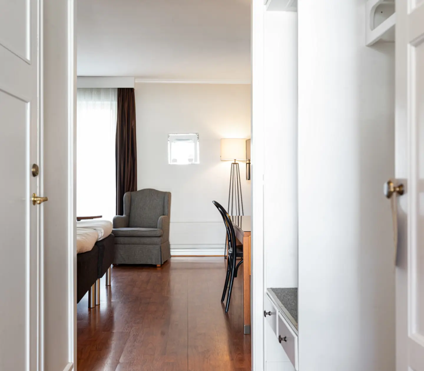 Modern hotel room with wooden flooring, featuring a cozy armchair, a neatly made bed, a desk with a chair, and a floor lamp, viewed from an open doorway.
