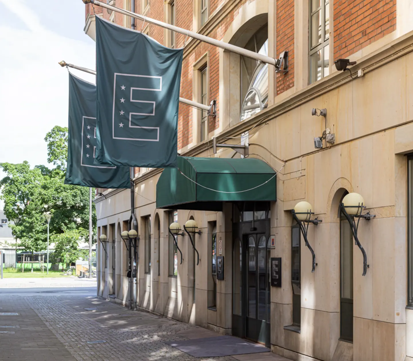 Facade of a European hotel with a green awning and flags displaying an 'E' logo, located on a cobblestone street.