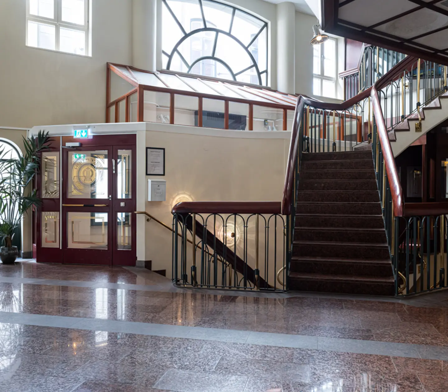Elegant hotel lobby with marble flooring, curved staircase, and large windows, featuring a decorative entrance door and potted plants.