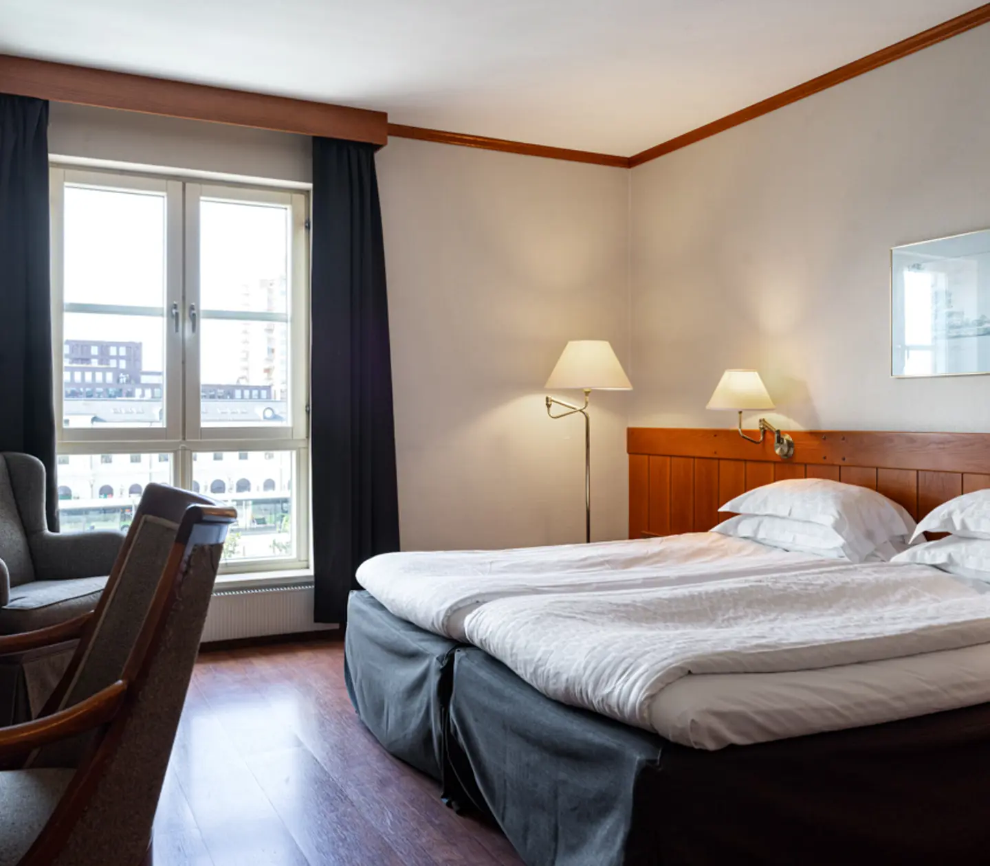 Modern hotel room with a large bed, two bedside lamps, a framed picture on the wall, and a chair near a window with city views.