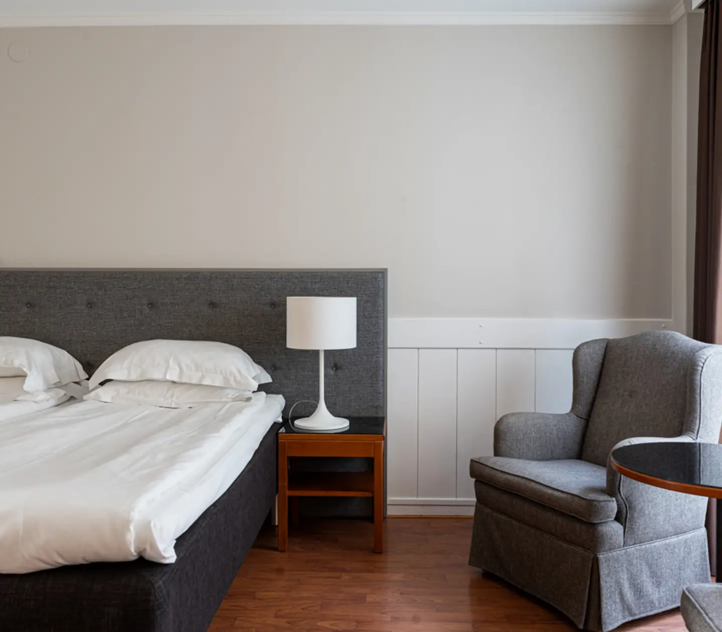 Modern hotel room with a neatly made double bed, gray upholstered headboard, bedside table with a lamp, and a comfortable gray armchair next to a round glass table, all set against a neutral-toned wall with natural light streaming in.