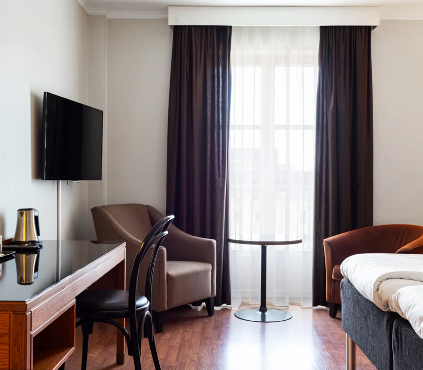 Hotel room interior featuring a comfortable bed, wooden desk with a chair, wall-mounted TV, and cozy seating area near a window with dark curtains.