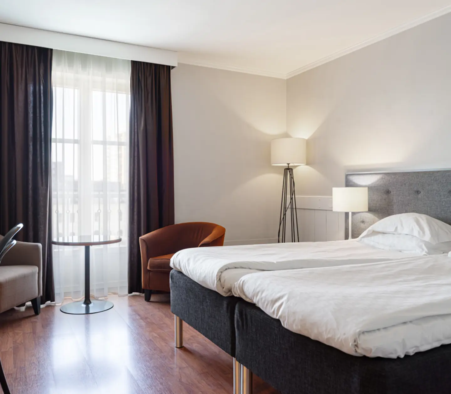 Modern hotel room with twin beds, stylish gray headboard, wooden flooring, floor lamp, and natural light from a window with brown curtains.