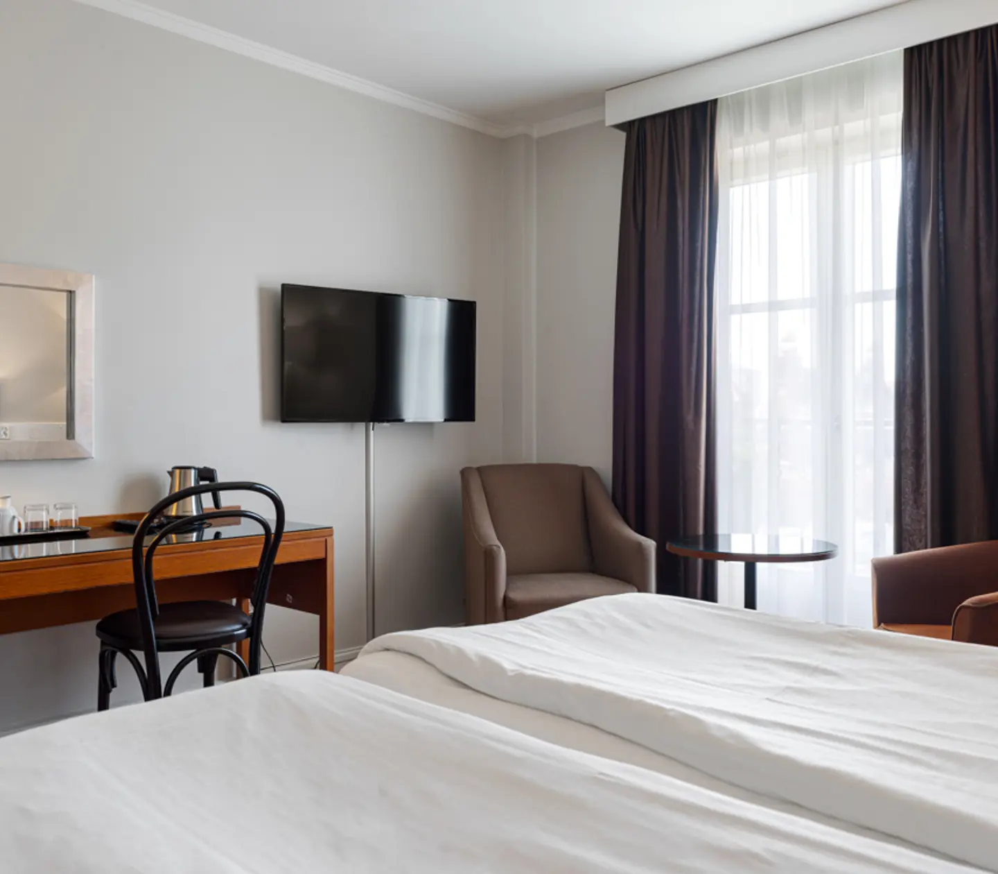 Modern hotel room with a comfortable bed, wall-mounted TV, desk with chair, armchair, small round table, and large window with brown curtains allowing natural light in.