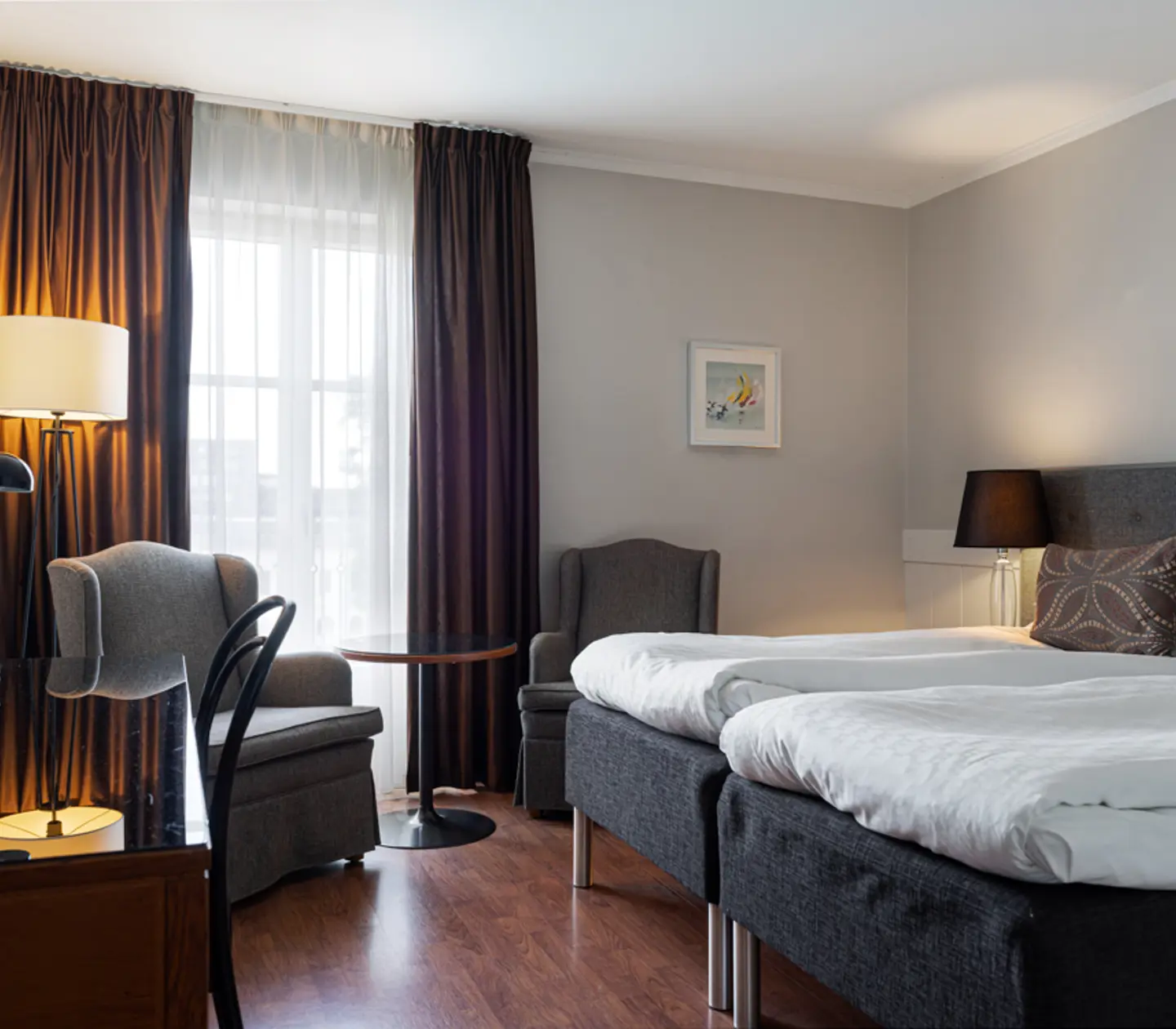 Cozy hotel room with two twin beds, modern furnishings, gray armchairs, a desk with a lamp, and a large window with curtains.