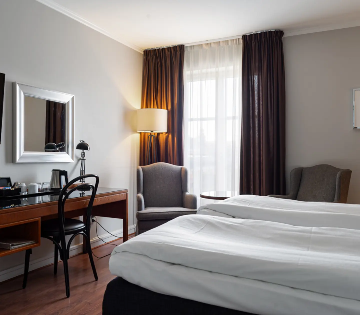 Hotel room with twin beds, a work desk, and two armchairs by the window with curtains. Cozy lighting from a floor lamp creates a relaxing atmosphere.