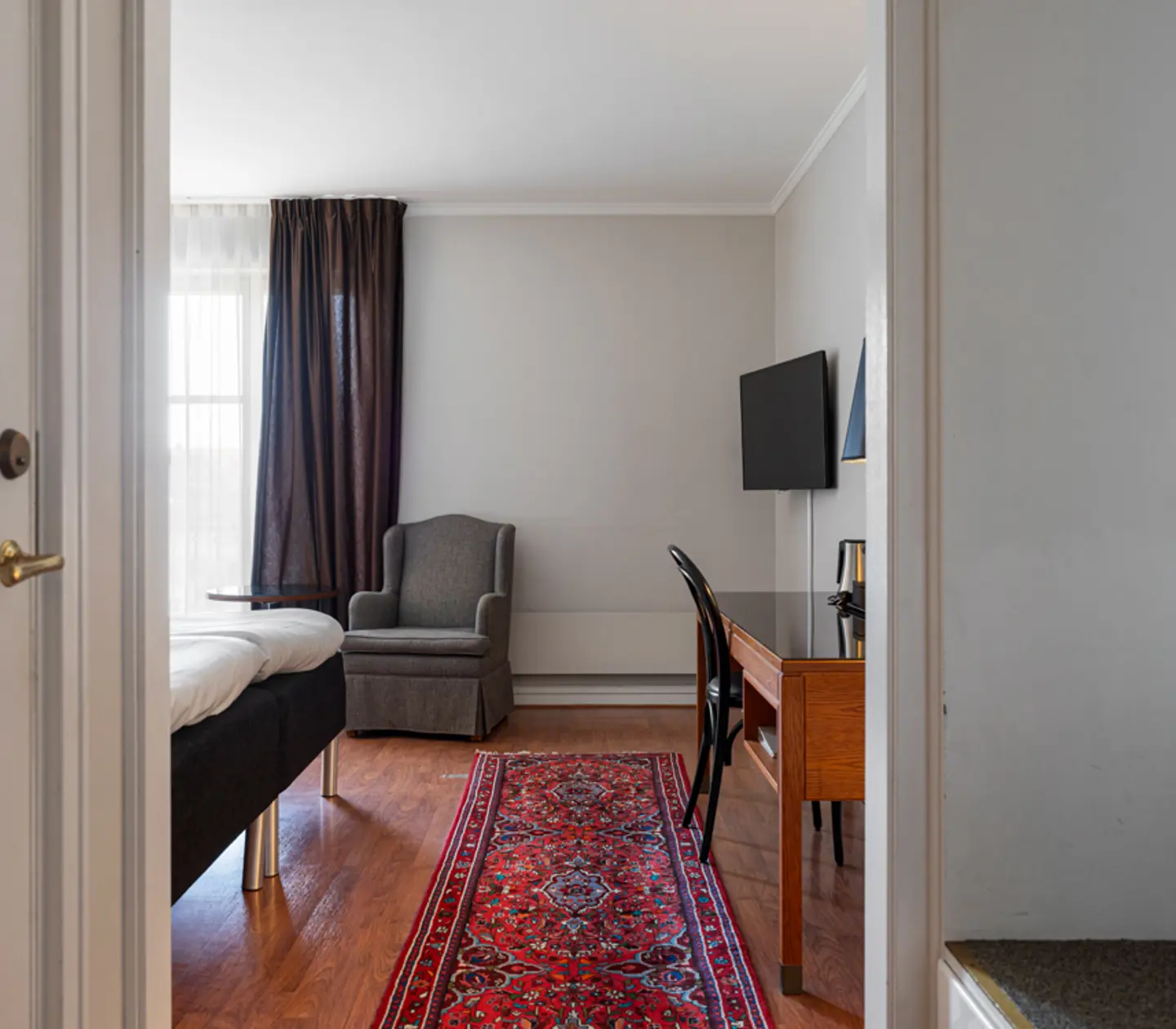 Hotel room with modern decor featuring a bed, armchair, desk with chair, flat-screen TV, and a vibrant red patterned rug on a wooden floor. Natural light filters through large curtains.