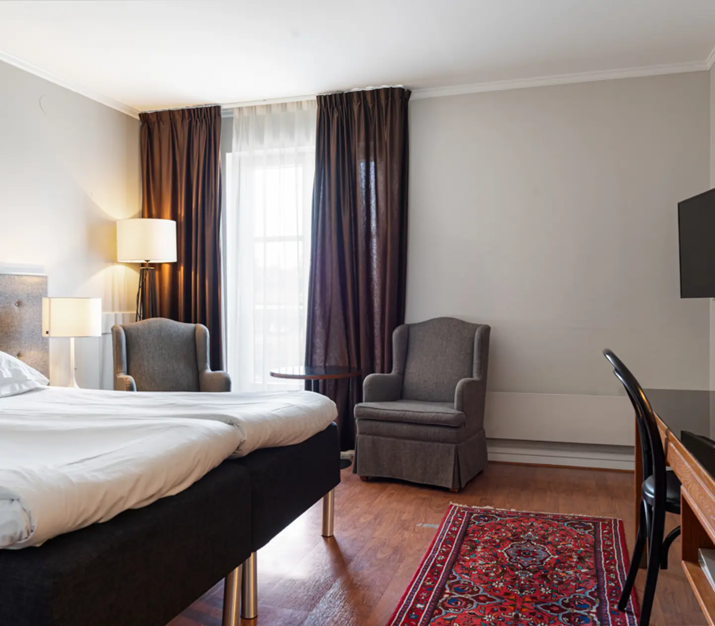 Modern hotel room with a comfortable bed, two upholstered armchairs, a flat-screen TV, and a wooden desk. The room features dark curtains, a large window, and a colorful rug.