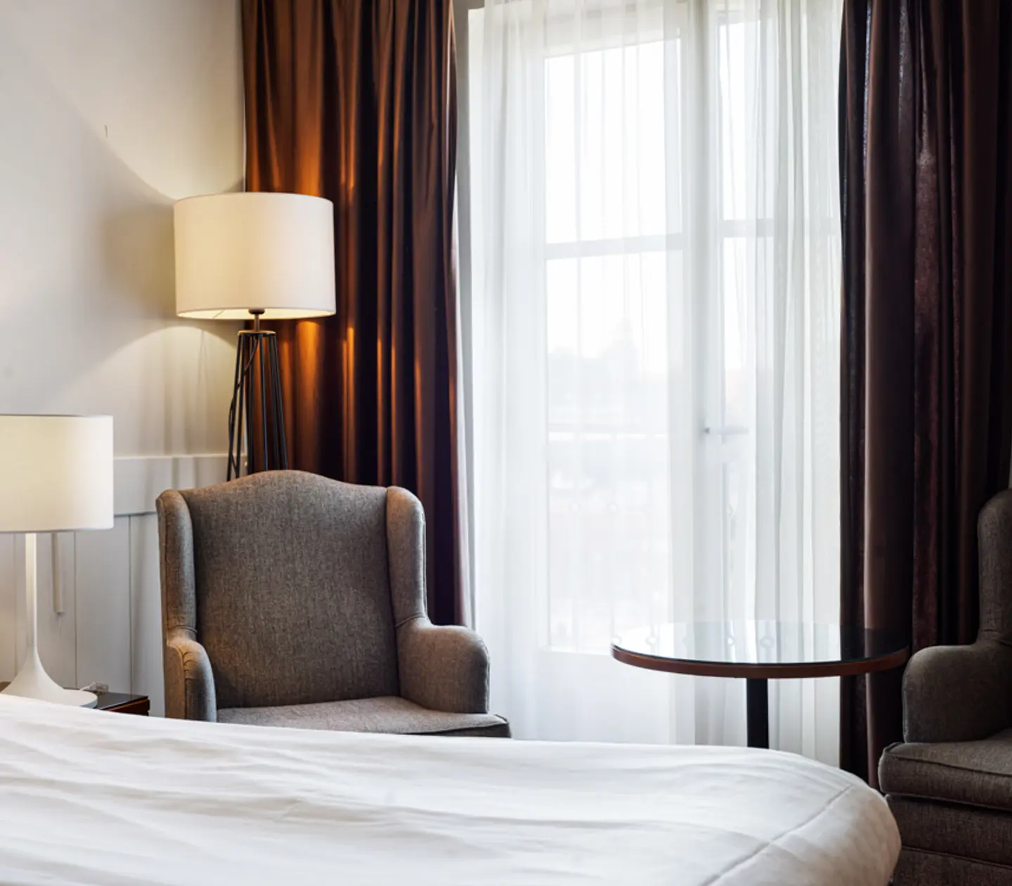 Cozy hotel room with two upholstered gray armchairs, bedside table, modern lamps, and sunlight filtering through sheer curtains.