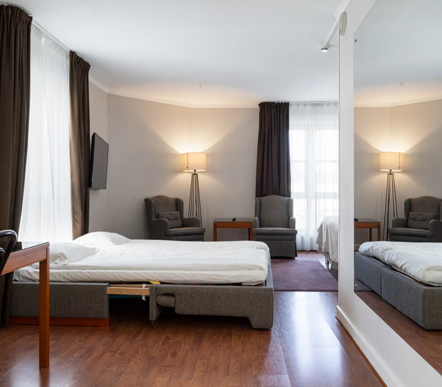 Contemporary hotel room with a single bed, cozy armchair, floor lamp, and large mirror reflecting the space. Room features neutral colors and wood flooring with natural light from large window.