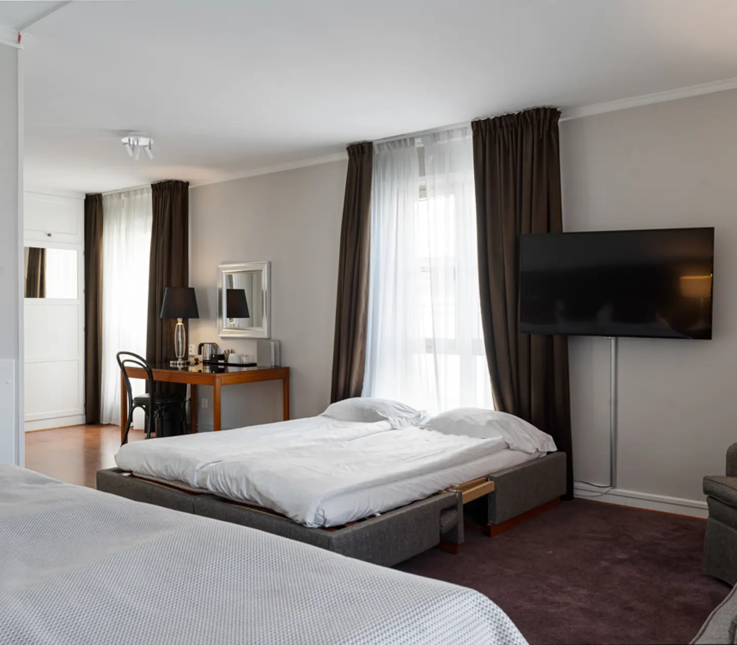 Modern hotel room with a double bed, sofa, large TV, and a desk by the window with dark curtains and wooden flooring.