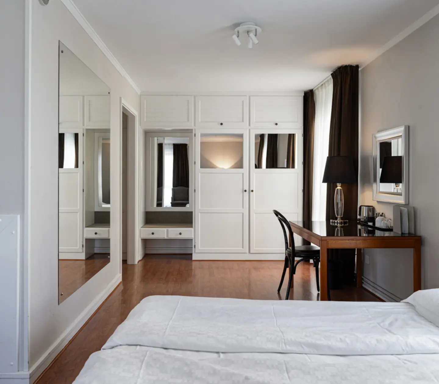 Modern hotel room with white bedding, large mirror, wooden desk, and wardrobe.