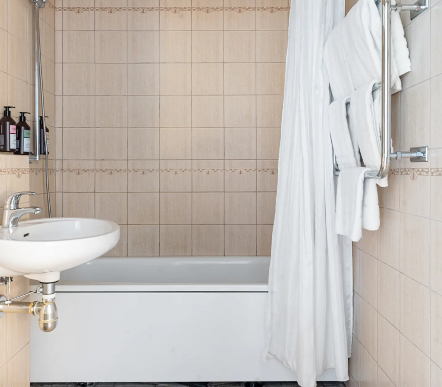 Modern bathroom with beige tiled walls, a white bathtub and sink, wall-mounted towel rack with white towels, and assorted toiletries on the bathtub edge.