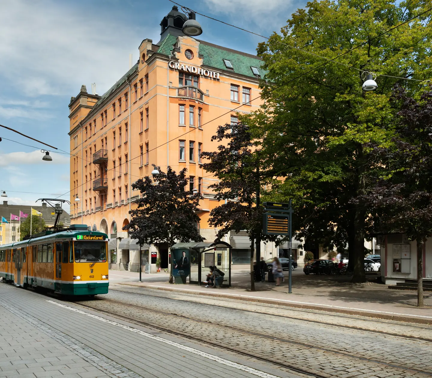 The orange facade of the Elite Grand Hotel in Norrköping with trees and a tram in front