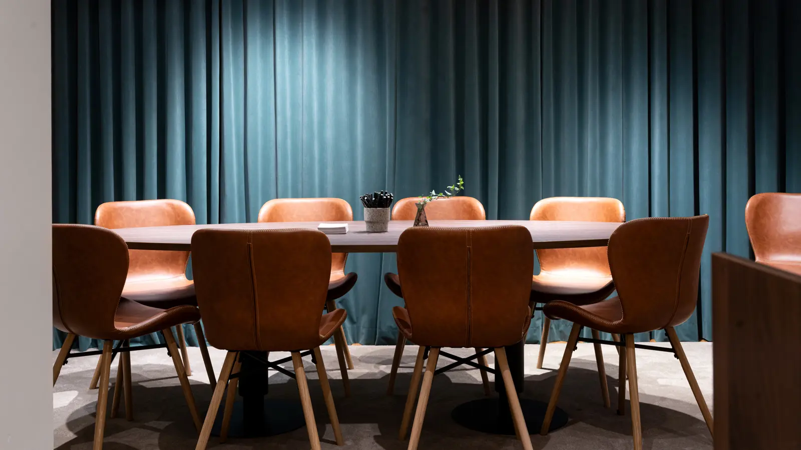 Modern conference room with a round table, surrounded by brown leather chairs, in front of dark teal curtains.