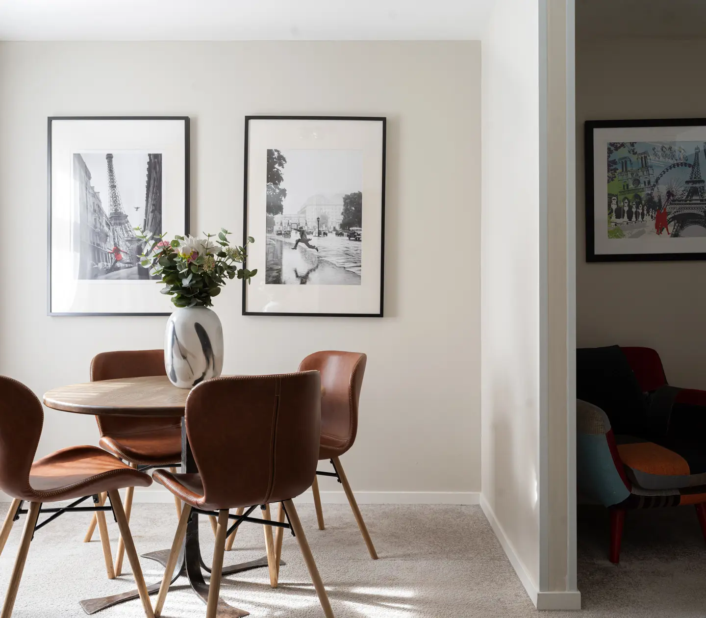 Modern dining area with round wooden table and brown leather chairs, featuring black and white artwork of Paris on the wall, complemented by a vase of flowers on the table.