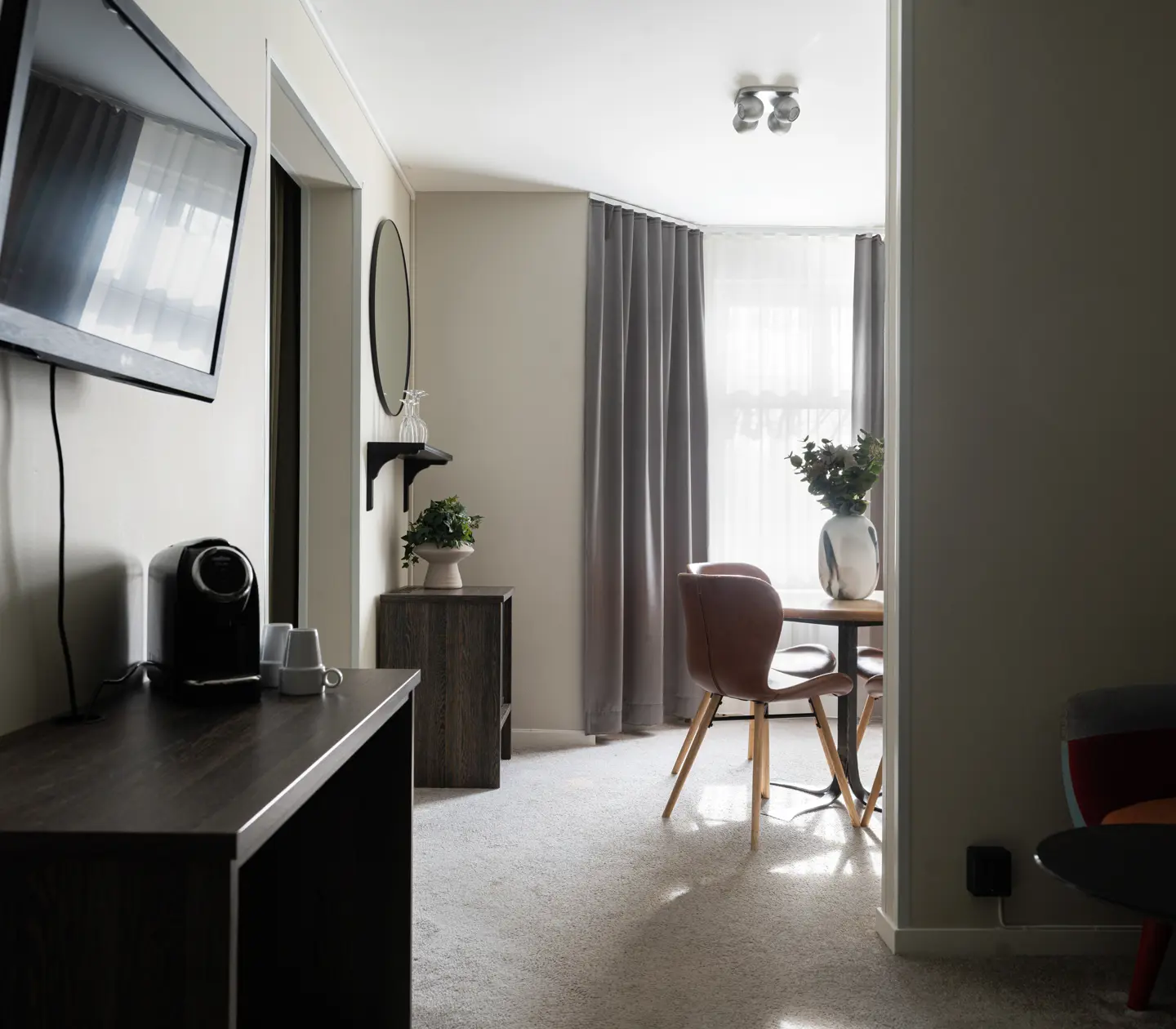 Modern living room with wall-mounted TV, coffee maker on a dark wood table, round dining table with brown chairs, large window with gray curtains, and vase with flowers on a side table. Bright and minimalistic interior design.