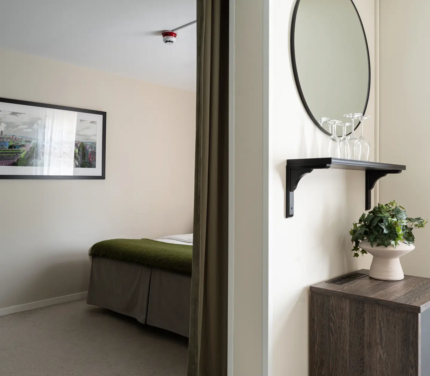 Modern bedroom interior with minimalist decor featuring a neatly made bed, a framed cityscape photo on the wall, and a wooden shelf with a mirror above it. The room has neutral tones and a potted plant for a touch of greenery.