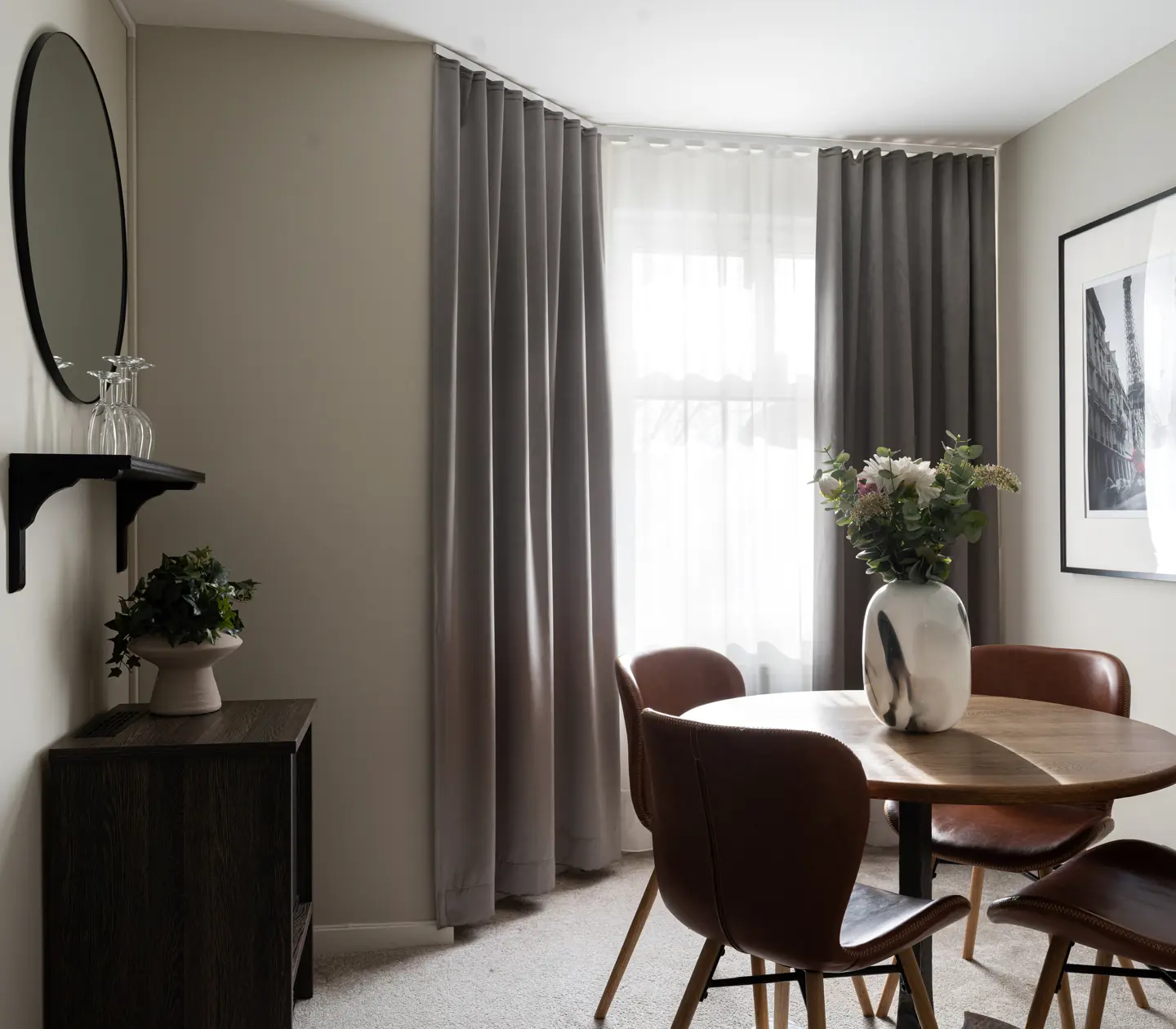 Modern dining area with round wooden table, brown leather chairs, and a vase of flowers. Soft gray curtains cover the window, and a framed photo adorns the wall. A wooden cabinet with plants and a circular mirror enhance the decor.