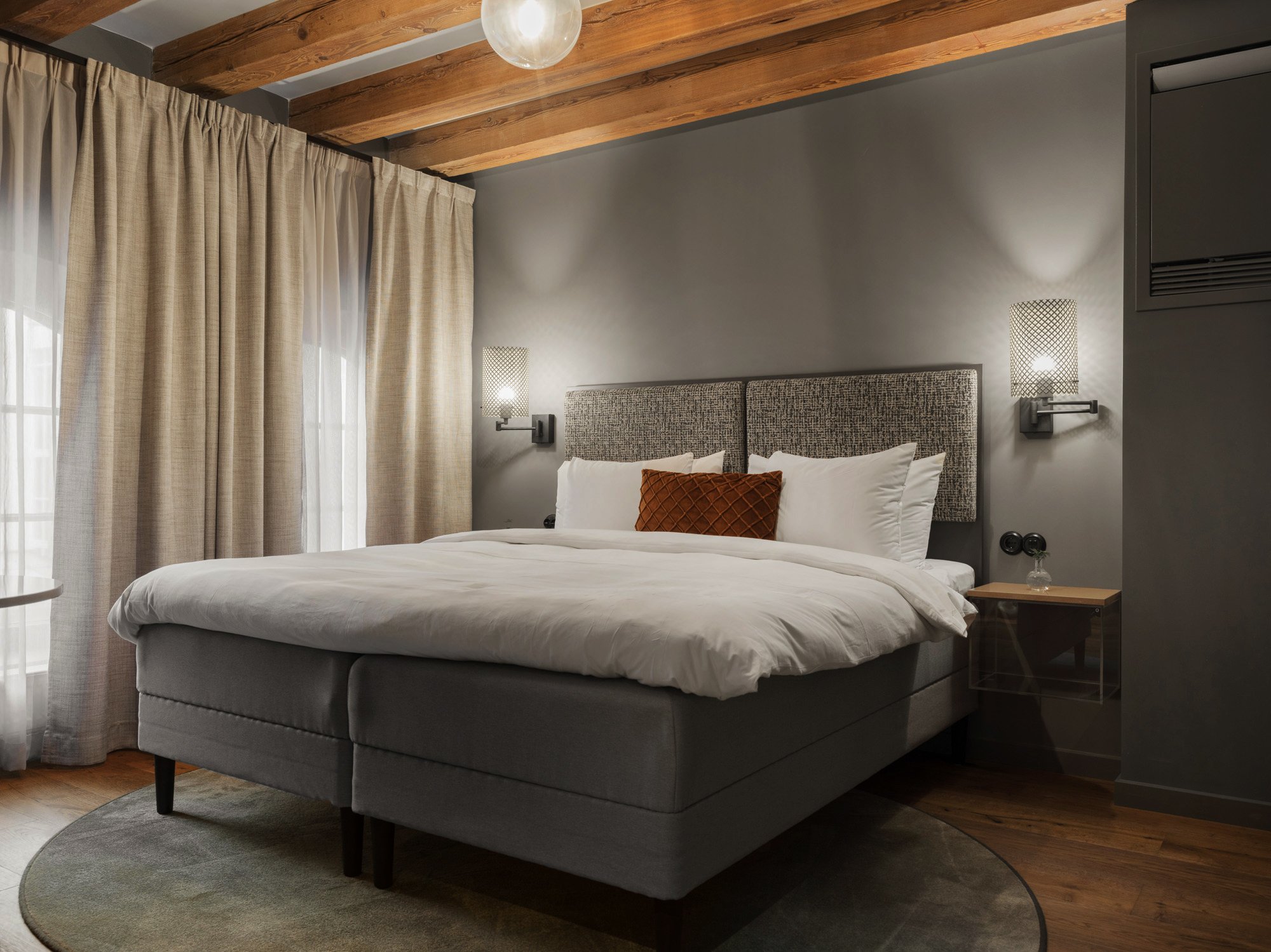 Ljust hotellrum med säng, beiga gardiner och träbalkar i taket