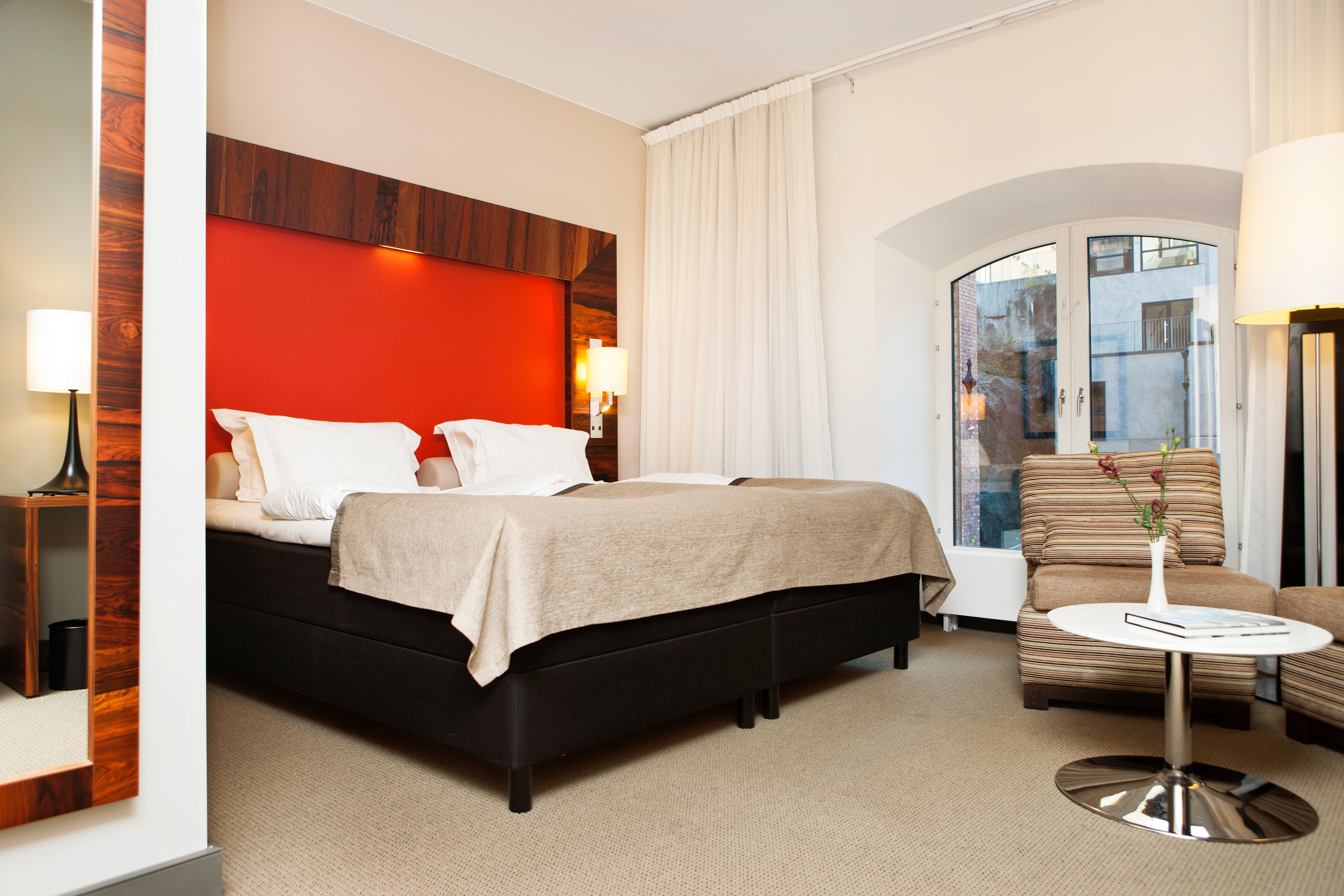 Ljust hotellrum med säng, röd sänggavel och fåtöljer