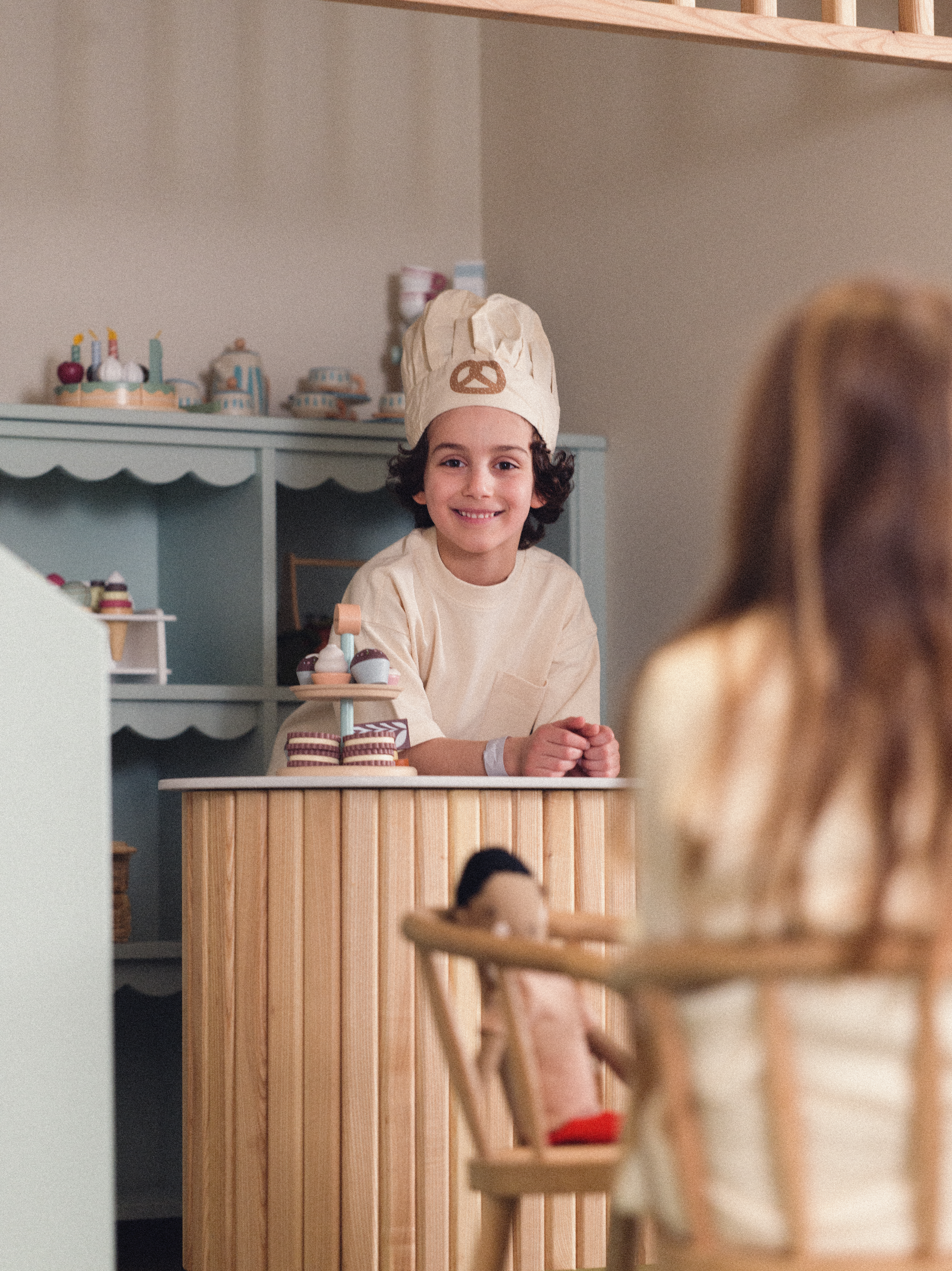 Child in chef costume behind a play kitchen counter, smiling, with play food and toys in a cozy playroom setting.