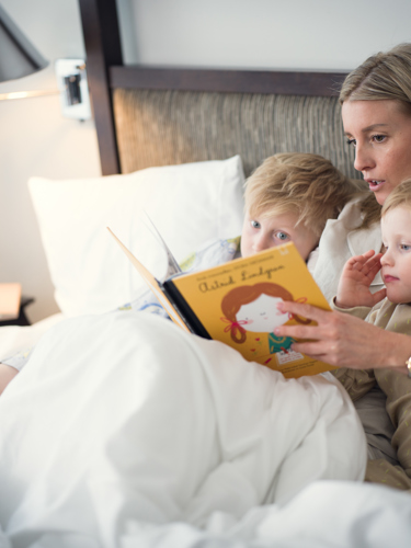 Mamma och två barn som sitter och läser bok i en hotellsäng