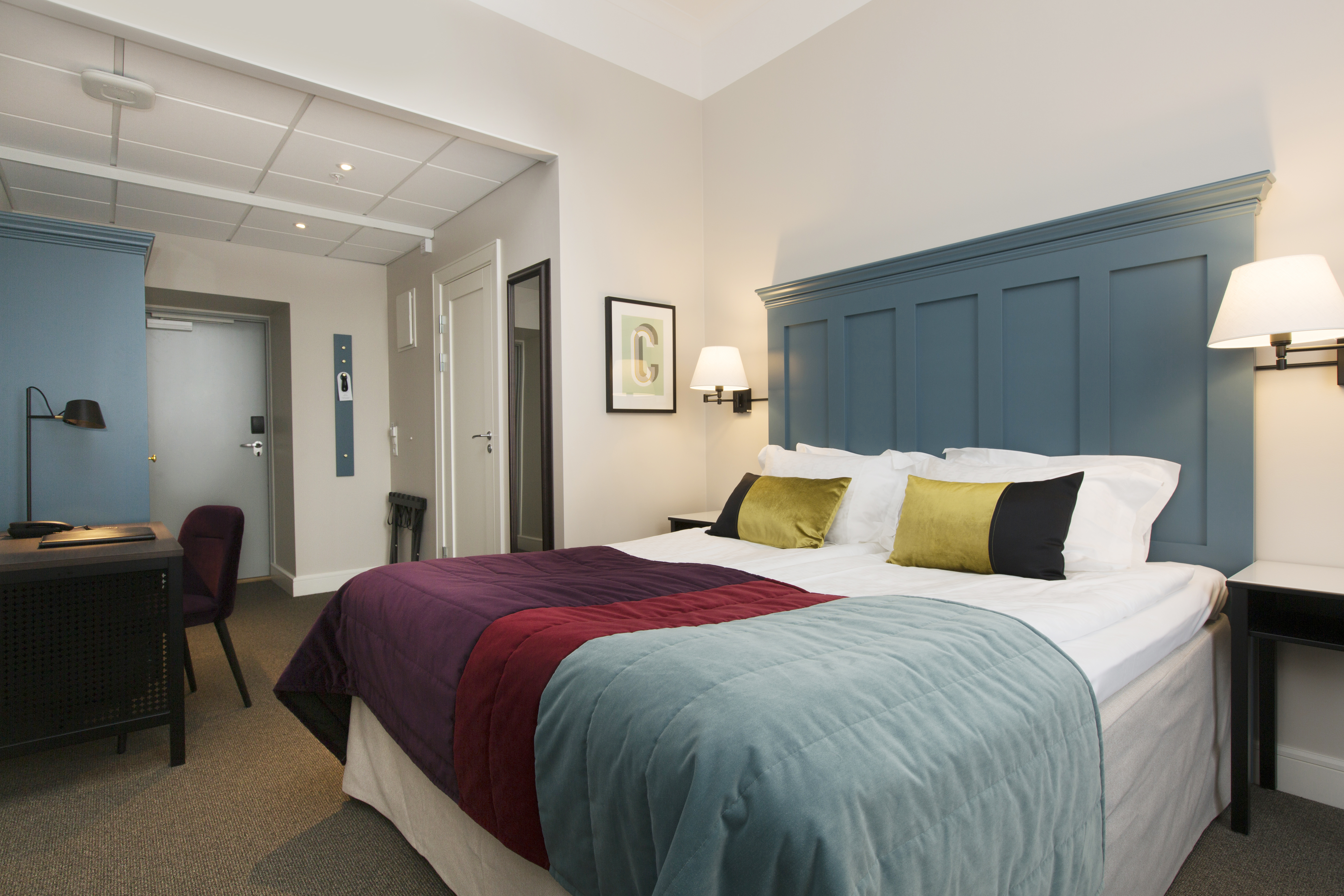 Ljust hotellrum med säng, stor blå sänggavel och skrivbord
