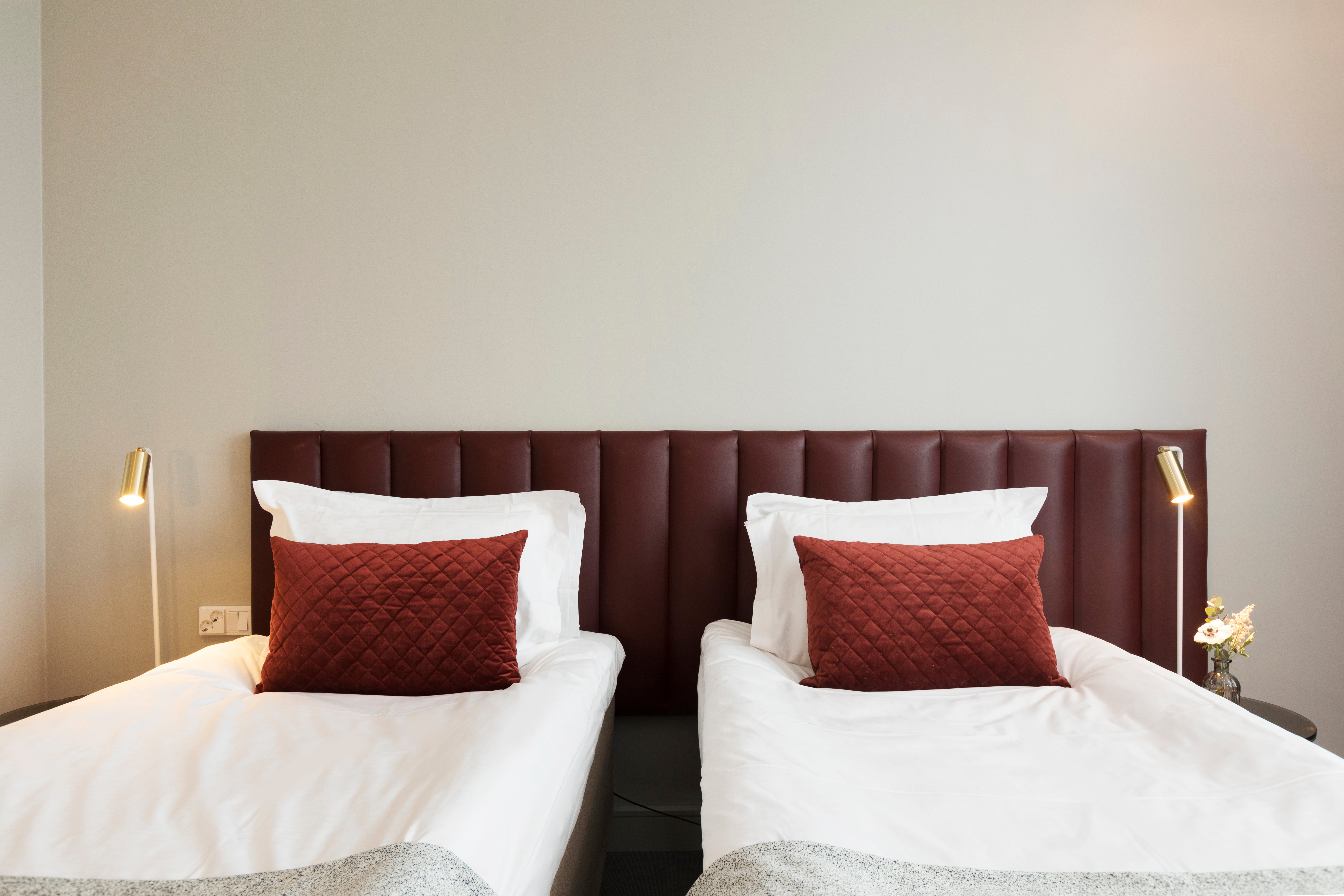 Ljust hotellrum med två enkelsängar bredvid varandra, vinröd sänggavel och små sänglampor