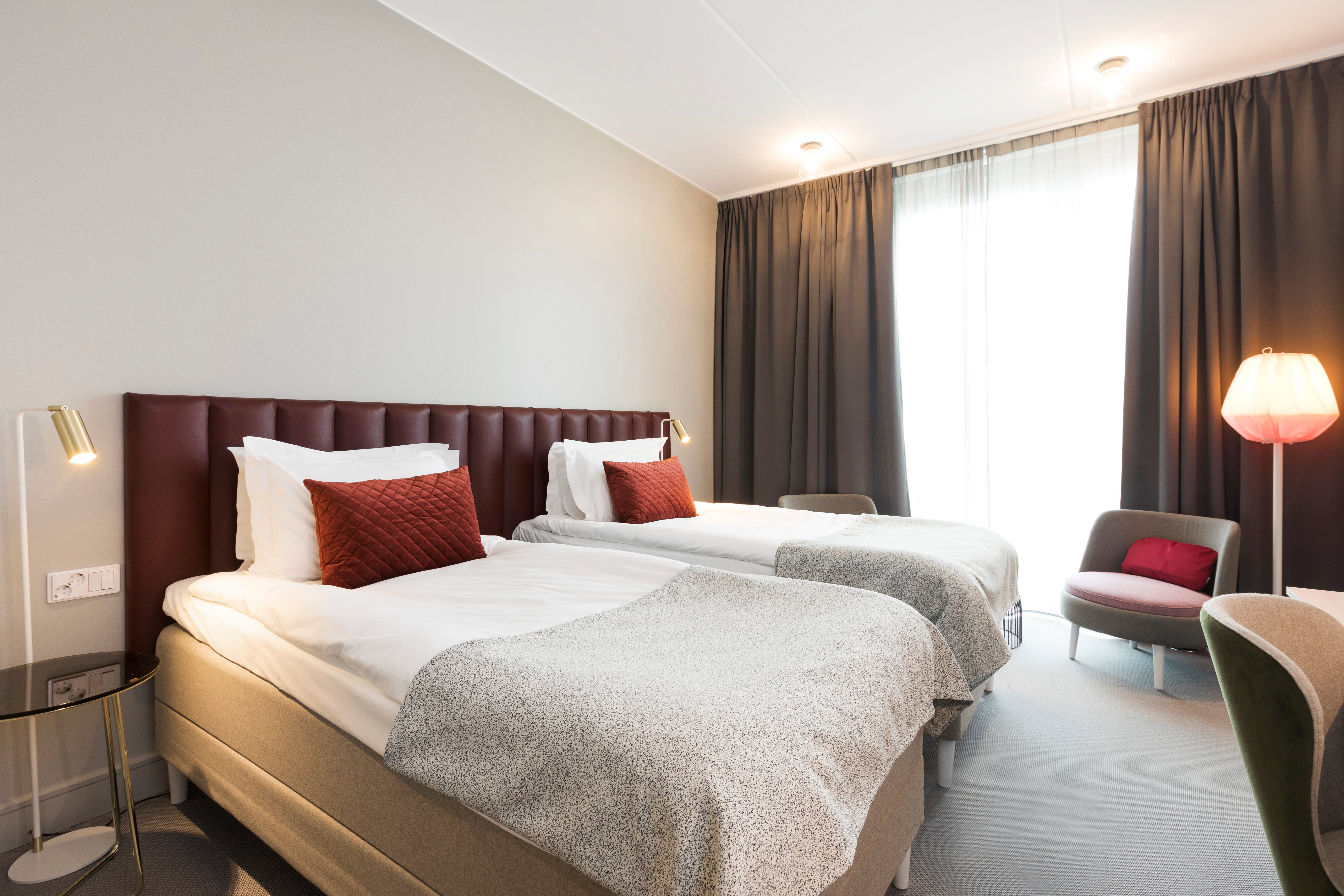 Ljust hotellrum med två enkelsängar bredvid varandra, vinröd sänggavel och fåtöljer
