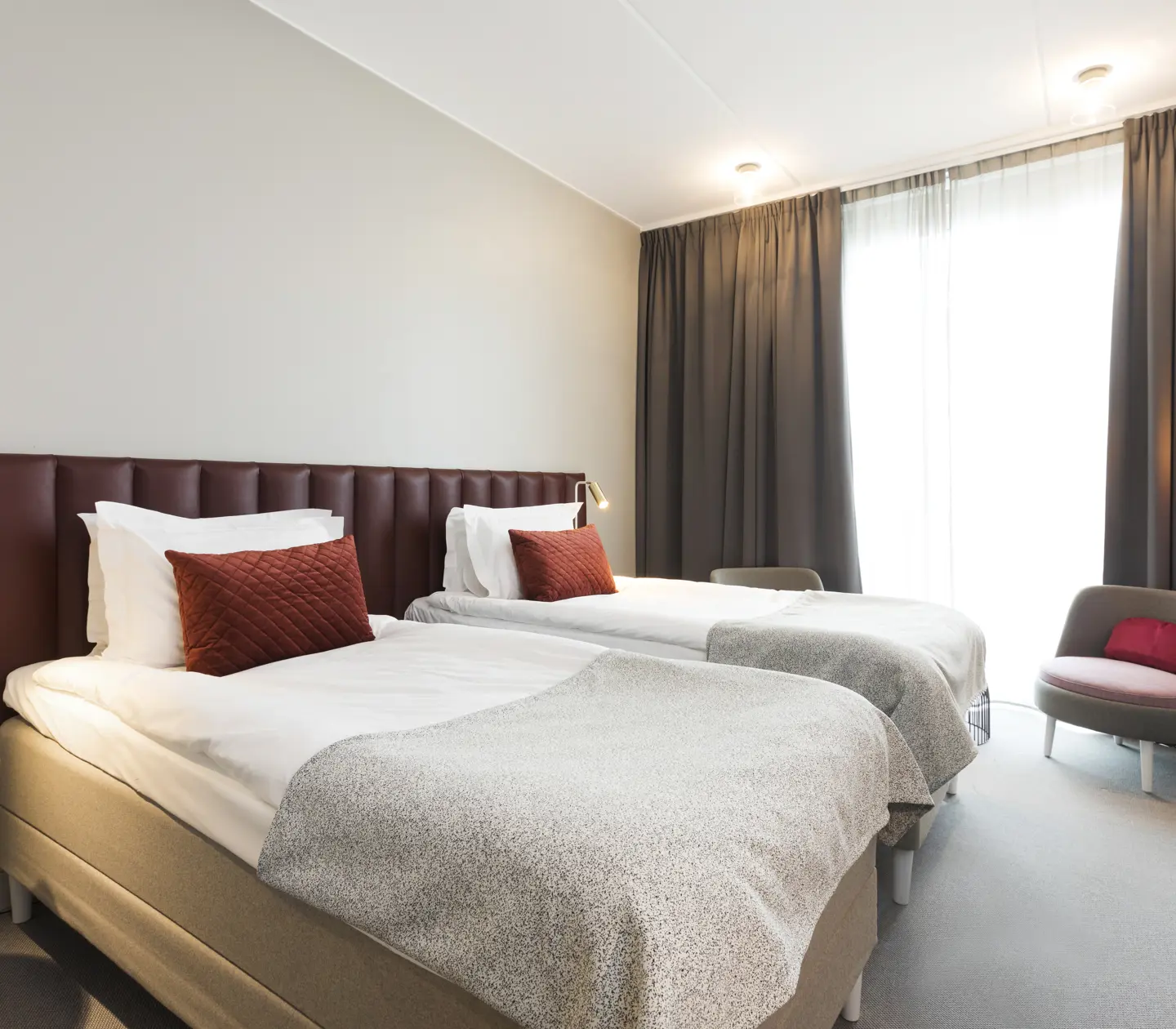 Bright hotel room with two single beds next to each other, burgundy headboard and armchairs