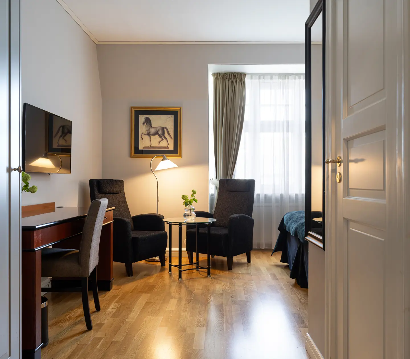 Cozy hotel room with wooden floors, featuring a desk, flat-screen TV, two armchairs, floor lamp, and a small table. A horse painting decorates the wall, and natural light filters through sheer curtains.
